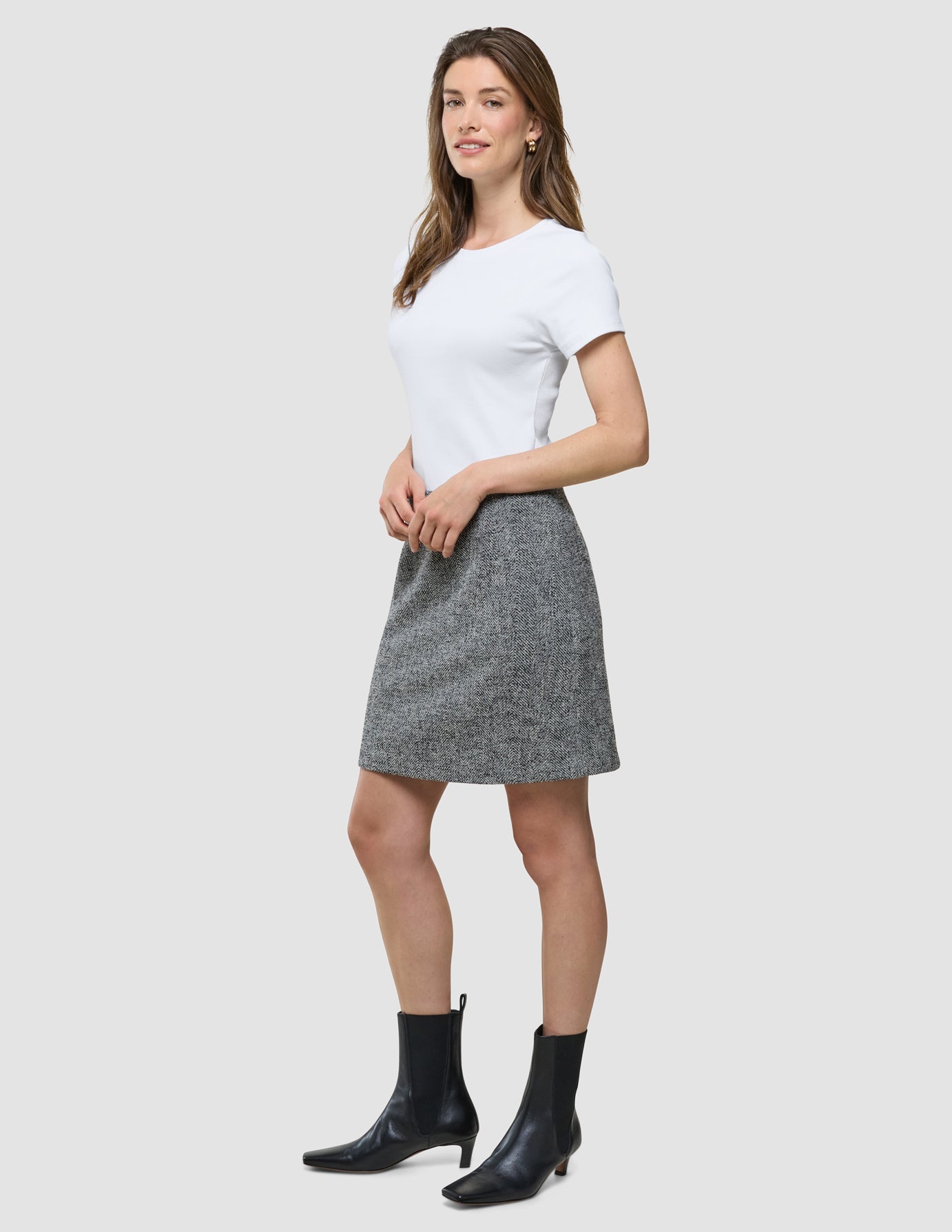 En kvinna bär en vit t-shirt, en kort kjol från Women Ashryn i mönstret ”Domino Herringbone” samt svarta ankelstövlar framför en enfärgad bakgrund.